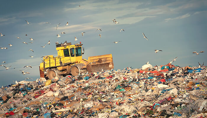 A yellow digger drives over trash at a landfill.