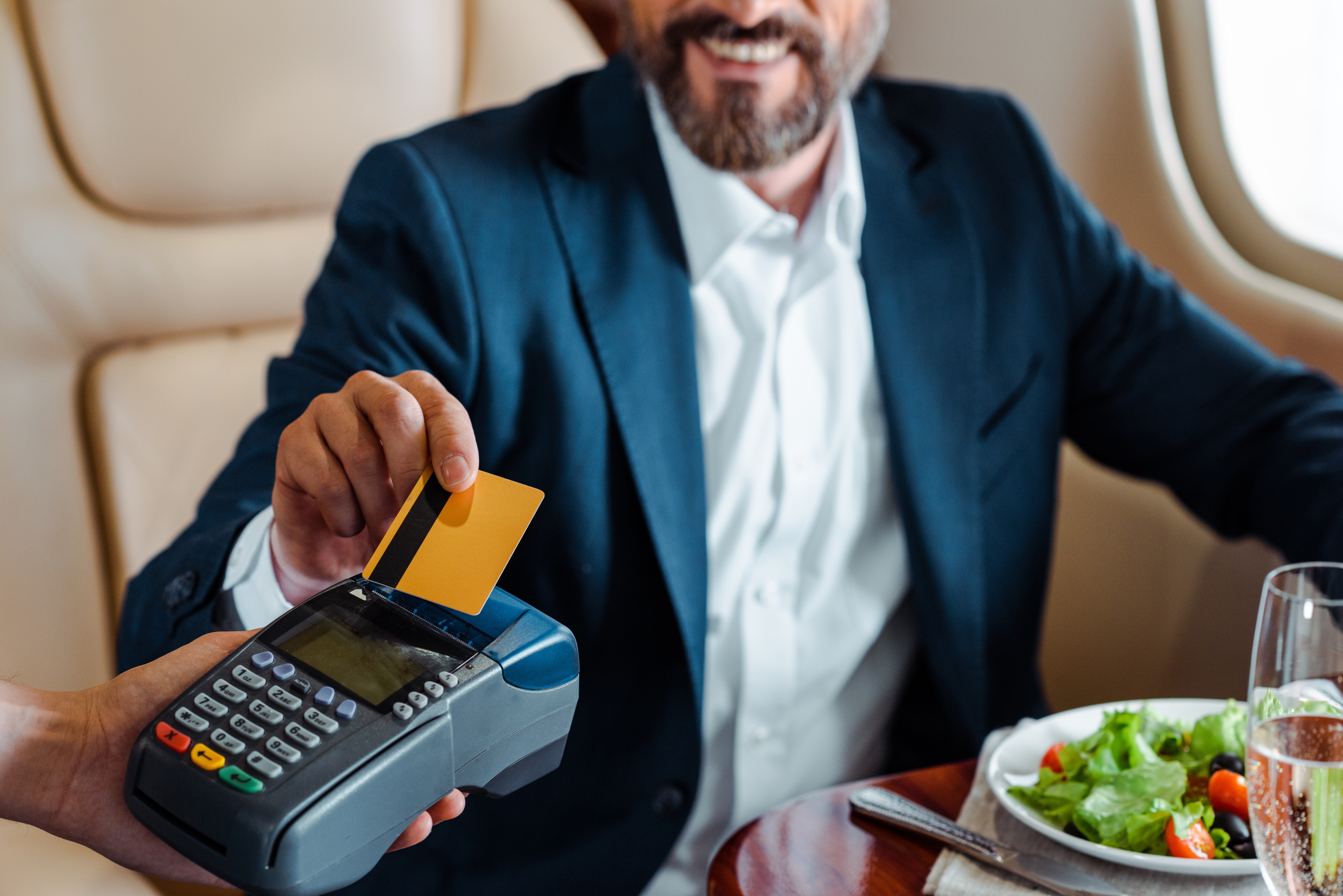 Selective focus of smiling businessman paying with credit card near salad and glass of champagne in airplane