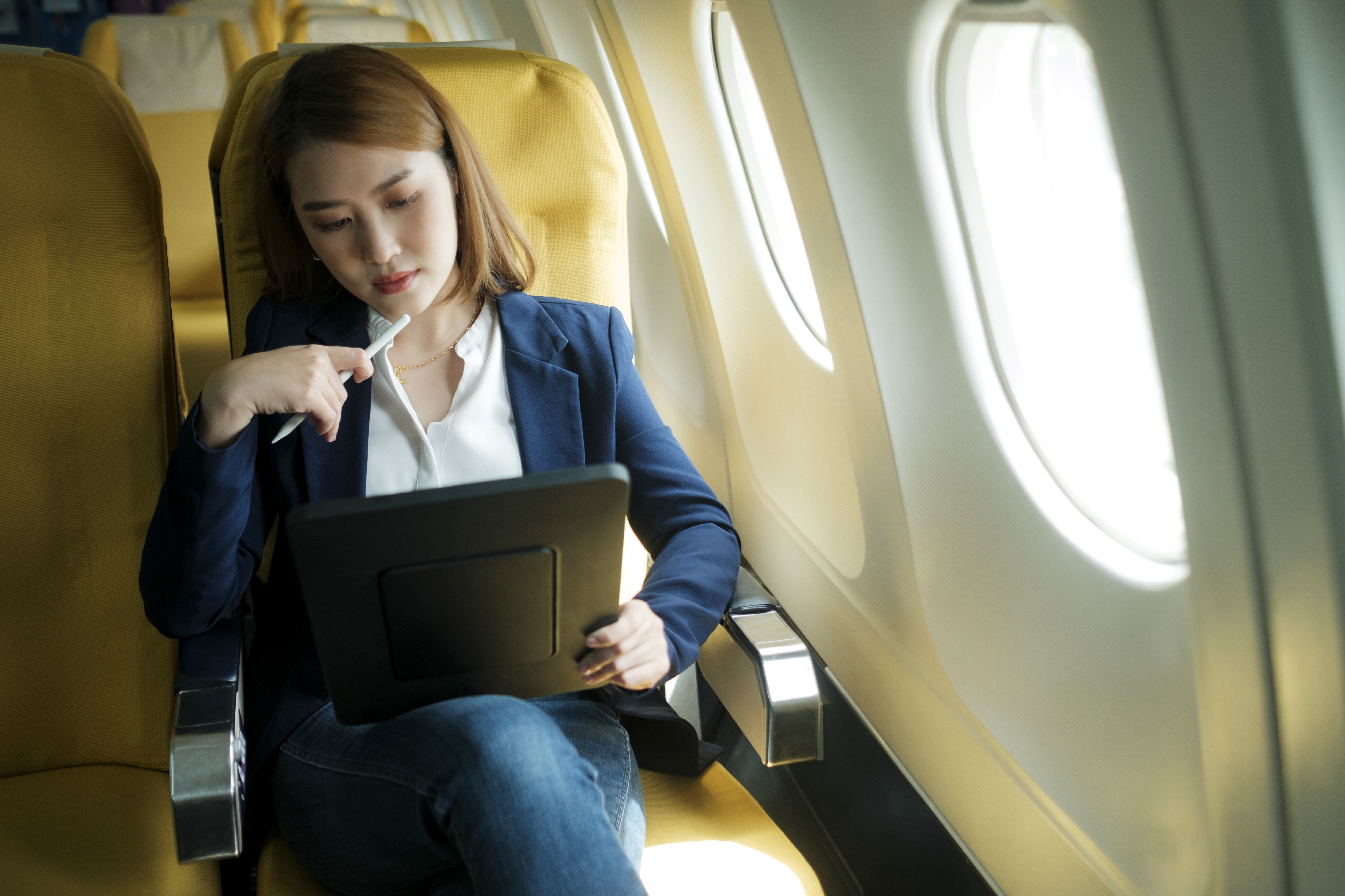 Young professional asian businesswoman in formal clothes working using tablet with smart pen while sitting in airplane cabin near window traveling to another place. Traveling and business concept.